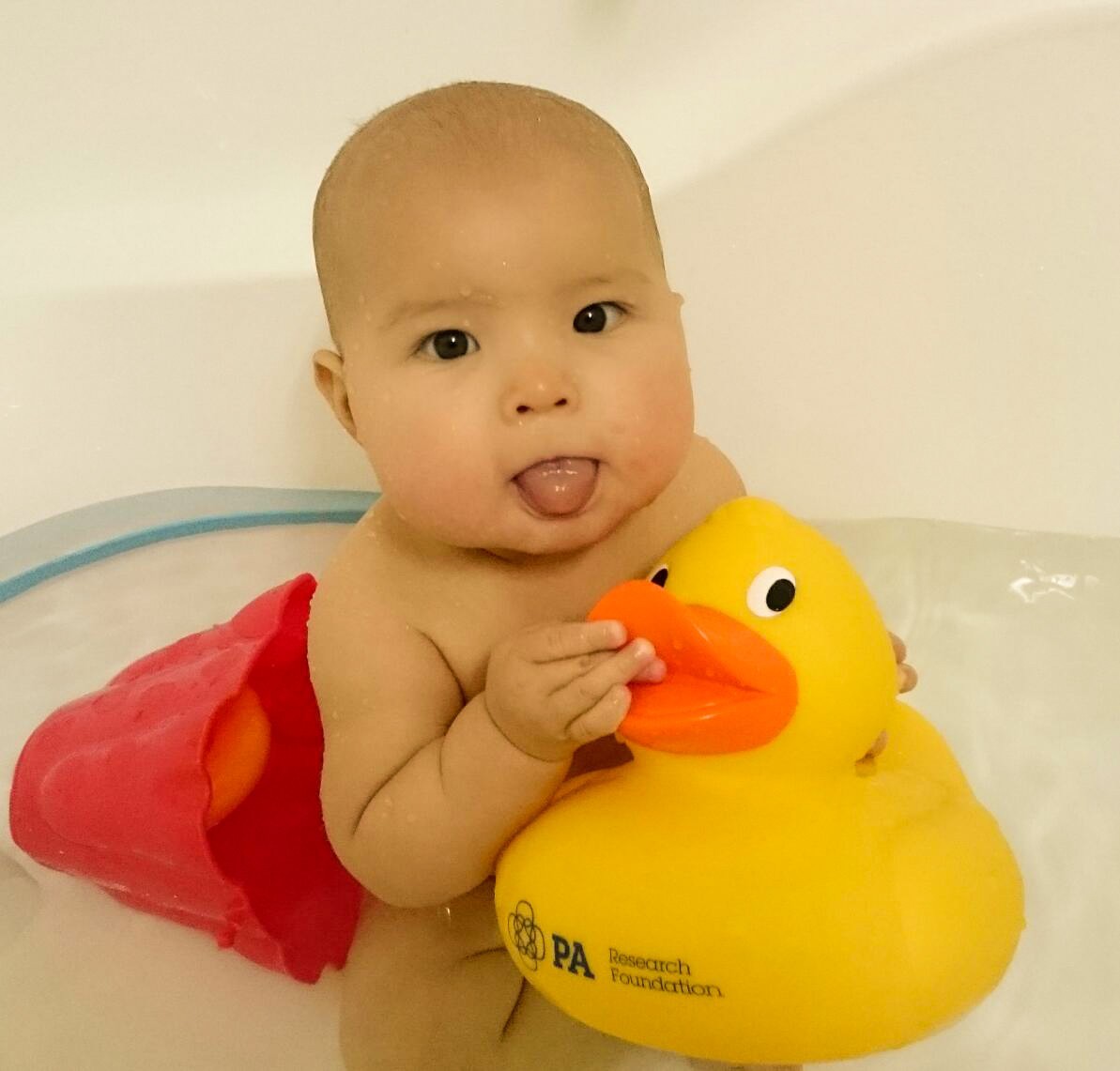 A smiling baby in the bath, sticking her tongue out, holding a rubber duck